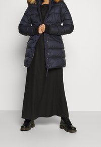Marineblauwe pufferjack met een met bont afgewerkte capuchon, voorzien van een gequilt ontwerp, ritssluiting en elastische manchetten. Draagt over een zwarte maxi-jurk.