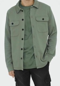 Chaqueta verde abotonada con dos bolsillos en el pecho, tela texturizada y botones negros, usada sobre una camisa verde a juego.