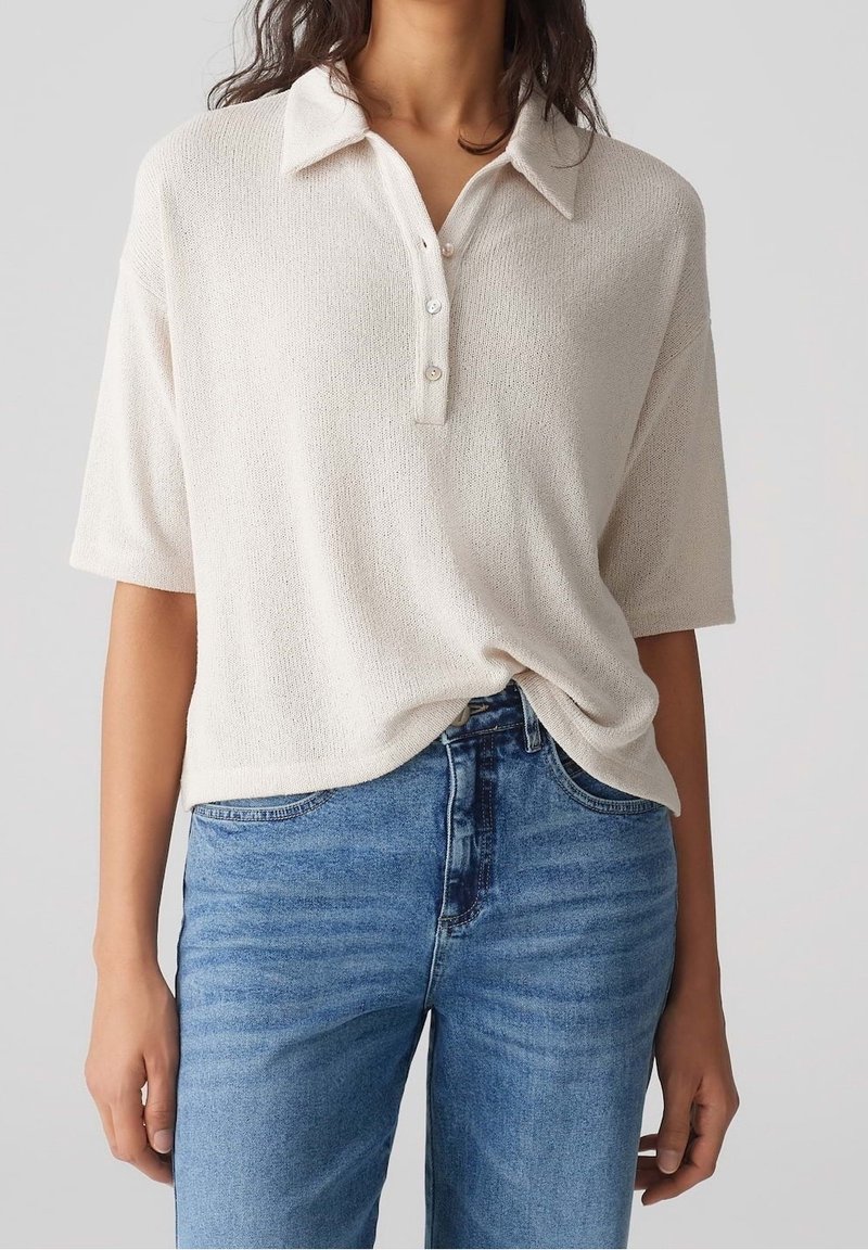 Ljusbeige kortärmad polo-tröja med strukturerad stickning, treknappsknäppning och en avslappnad passform, tillsammans med medelblå jeans.