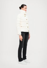 Giubbotto imbottito color crema, con colletto alto, bottoni dorati e imbottitura orizzontale. Abbinato a jeans neri e sneakers bianche.