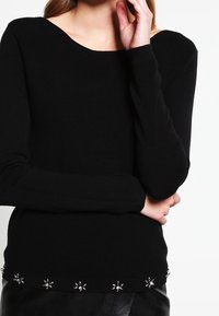 Femme portant un pull noir à manches longues avec un col asymétrique et des embellissements floraux en argent décoratifs le long de l'ourlet.