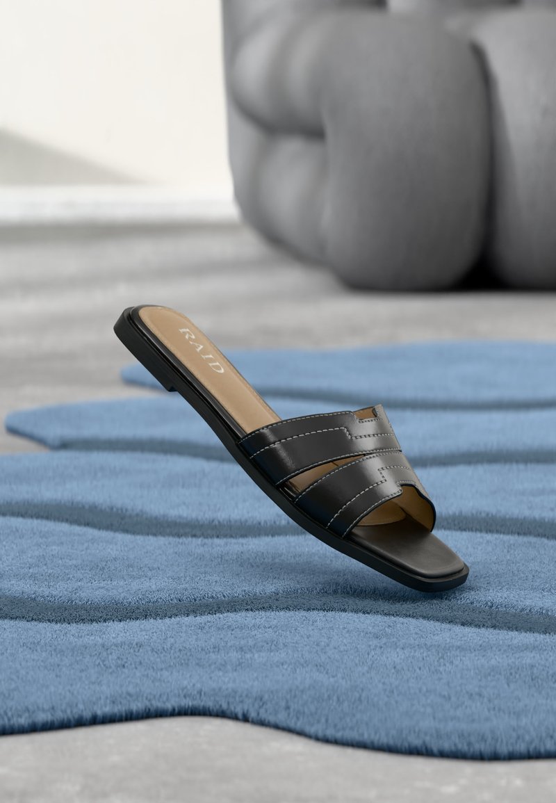 Sandale plate noire à enfiler avec larges brides cousues sur un tapis bleu texturé, avec un objet de siège abstrait flou en arrière-plan.