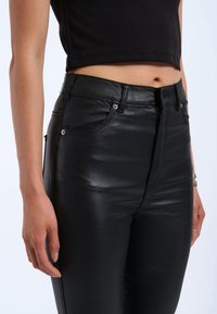 Zwarte faux leren high-waisted broeken met een aansluitend ontwerp, voorzien van voorzakken, metalen accenten en een gladde, glanzende textuur.