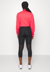 Dlhý rukáv, textúrovaný, jasne ružový top v kombinácii s priliehavými čiernymi legínami s priezračnými panelmi, končiacimi nad členkom. Biele a ružové tenisky.