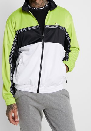Homme portant une veste de survêtement FILA vert lime, noir et blanc par-dessus un t-shirt noir et un pantalon de survêtement gris, avec une main dans la poche et des tatouages visibles.