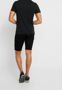 Hombre con camiseta negra, pantalones cortos negros y zapatillas negras de pie de espaldas a la cámara frente a un fondo blanco.
