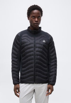 Man wearing a black quilted jacket with ACG logo on chest, standing against a plain light background, hands relaxed by sides.