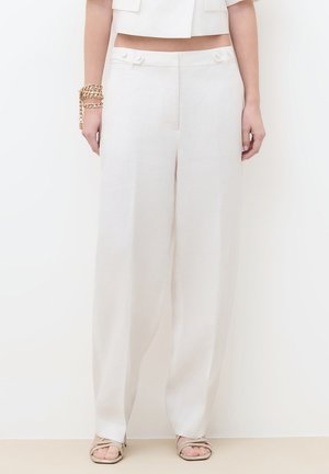 Kobieta w białych, szerokich spodniach z wysokim stanem, do których dobrano krótki biały top, złotą bransoletkę z łańcuszkiem oraz złote sandały na otwartym palcu.