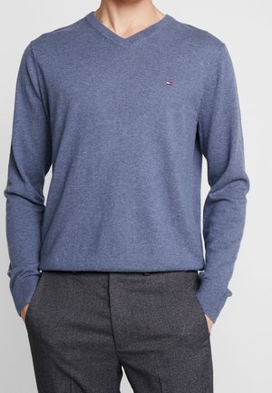 Mand iført en blå V-hals sweater med et lille logo på brystet og mørkegrå bukser, hænderne i lommerne.