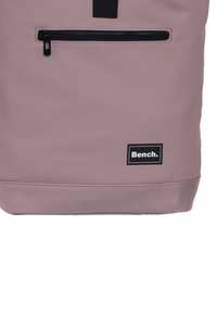 Paarse nylon rugzak met een voorzak met rits, zwarte hardware en een witte "Bench." logo op een zwart label nabij de basis.