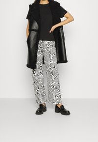 Chaleco sin mangas de cuero sintético negro sobre una camiseta negra, combinado con pantalones blancos que presentan un patrón abstracto negro y zapatos negros voluminosos.