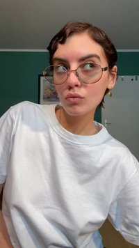 Okrogle očala s srebrnimi poudarki, bela prevelika majica, kratki temni lasje z mehko valovitostjo in zlata uhan v obliki kapljice.