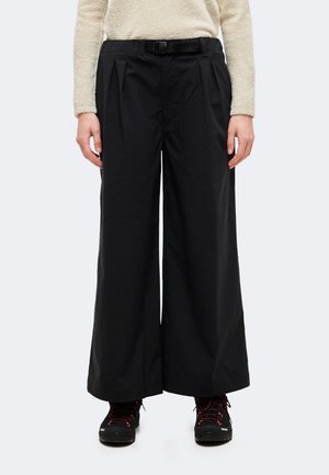 Une personne porte un pantalon noir à jambes larges avec une ceinture, un haut en polaire couleur crème et des chaussures de randonnée noires à lacets rouges.