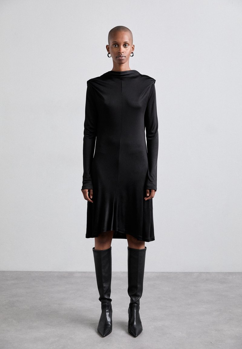 Black long-sleeve dress with a hood, smooth texture, knee-length hem, and slim fit. Paired with knee-high pointy-toed boots.