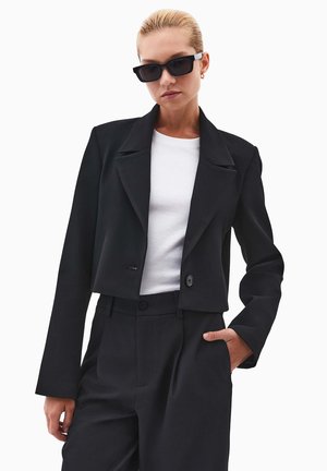 Blazer nero tagliato con ampi rever, due bottoni e una texture di tessuto liscia, abbinato a pantaloni neri a vita alta e a una maglietta bianca.