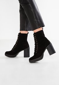 Bottes basses en daim noir avec un talon épais et une plateforme, dotées d'œillets dorés, d'un laçage à l'avant et d'un talon texturé.
