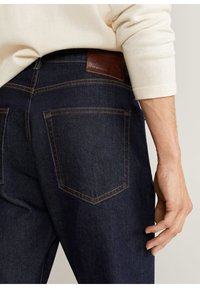Jeans in denim scuro con una toppa in pelle marrone in vita, cuciture in rilievo e due tasche posteriori visibili, tenuti da una mano.
