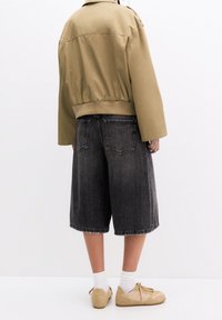 Person iført en kort tan jakke, løse sorte denimshorts i knæhøjde, hvide sokker og beige sneakers, stående mod en simpel hvid baggrund.
