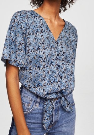 Blå mønstret bluse med V-hals, korte ærmer og bindebånd ved taljen. Har blomstermønster i forskellige nuancer af blå og hvid.