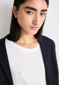 Primo piano di una persona che indossa un blazer blu scuro sopra una maglietta bianca, con texture lisce, scollatura rotonda e un design minimalista.
