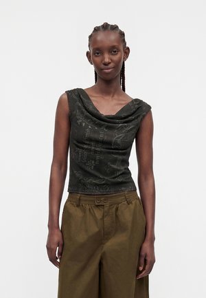 Femme aux cheveux tressés portant un haut noir sans manches à motifs et un pantalon ample vert olive, debout devant un fond uni.