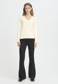 Krémový sveter s výstrihom do V z mäkkej pleteniny, kombinovaný s čiernymi zvonovými nohavicami. Sveter má dlhé rukávy a rebrované lemy; nohavice majú voľný strih.