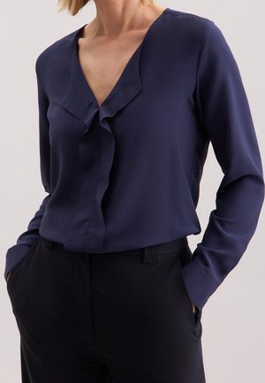 Kvinde iført en langærmet navybluse med flæser ved V-udskæringen, stoppet ned i sorte bukser, hænder delvist i lommerne.