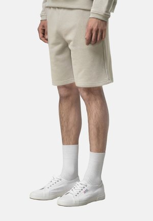 Uomo con pantaloncini beige, calzini bianchi e scarpe da ginnastica bianche in tela, in piedi davanti a uno sfondo semplice.