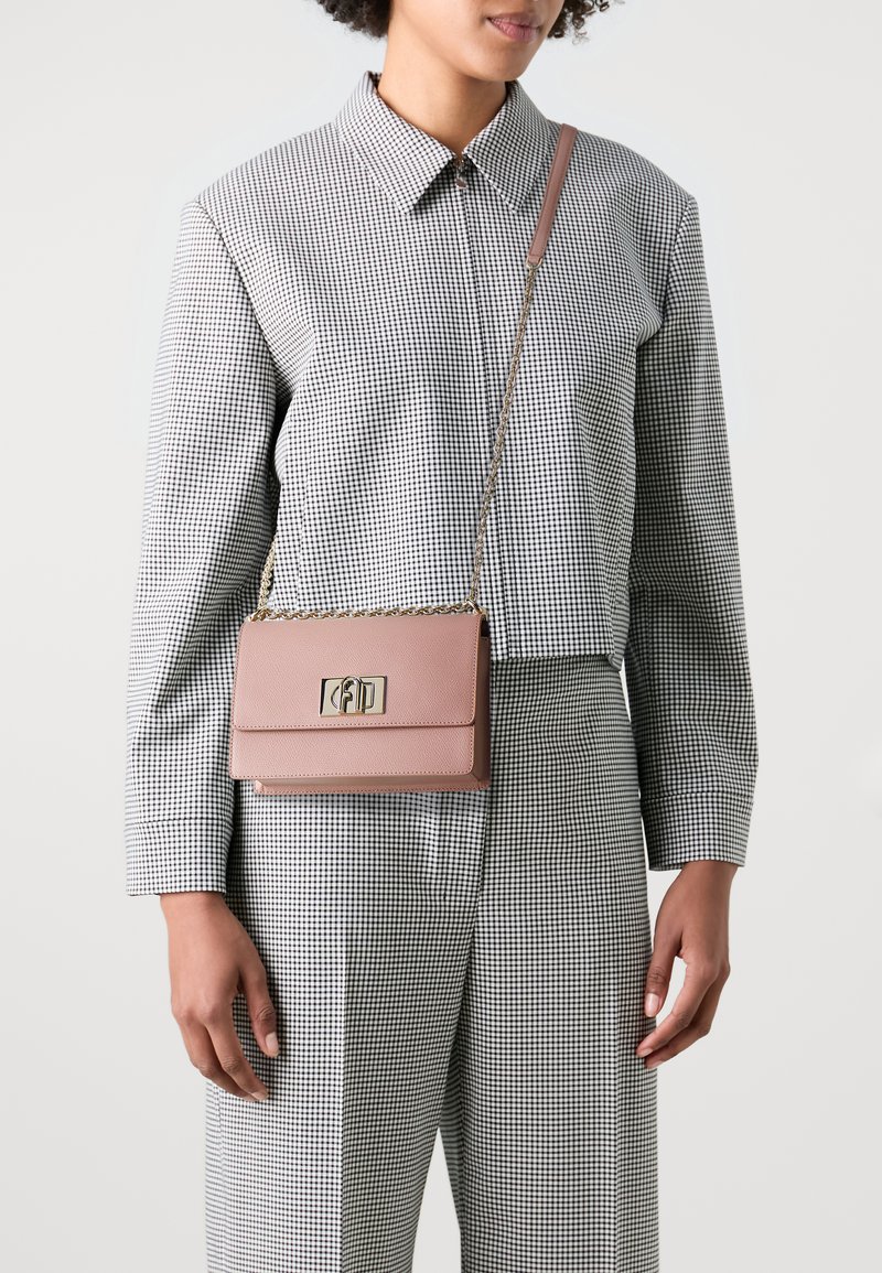 Borsa a tracolla in pelle rosa con hardware dorato, forma rettangolare e tracolla a catena. Indossata da una persona con un outfit a quadri.