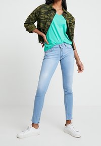 Chaqueta verde con patrón de camuflaje, camiseta turquesa, jeans ajustados de color azul claro y zapatillas blancas. Enfocado en ropa casual y ajustada.