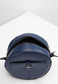 Borsa rotonda blu navy realizzata in pelle liscia, con chiusura a zip e una tasca esterna con chiusura a scatto, design minimalista.