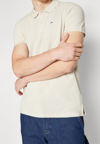 Mann in beige Polo-Shirt mit kurzen Ärmeln und blauen Jeans, mit verschränkten Armen, bis zur Hals- und Taillenlinie zugeschnitten, vor weißen Hintergrund stehend.