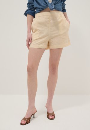 Frau trägt gelbe, strukturierte Shorts mit vorderen Taschen, ein blaues, vorne gebundenes Jeanshemd und braune Sandalen mit Absatz, sie steht vor einem einfarbigen Hintergrund.