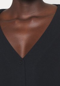 Närbild av en mörkhyad persons hals och övre bröst som bär en svart V-ringad ribbad topp med knappar.