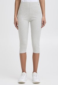 Personne portant des leggings cropped à côtes gris clair et des baskets blanches, se tenant devant un fond blanc uni.