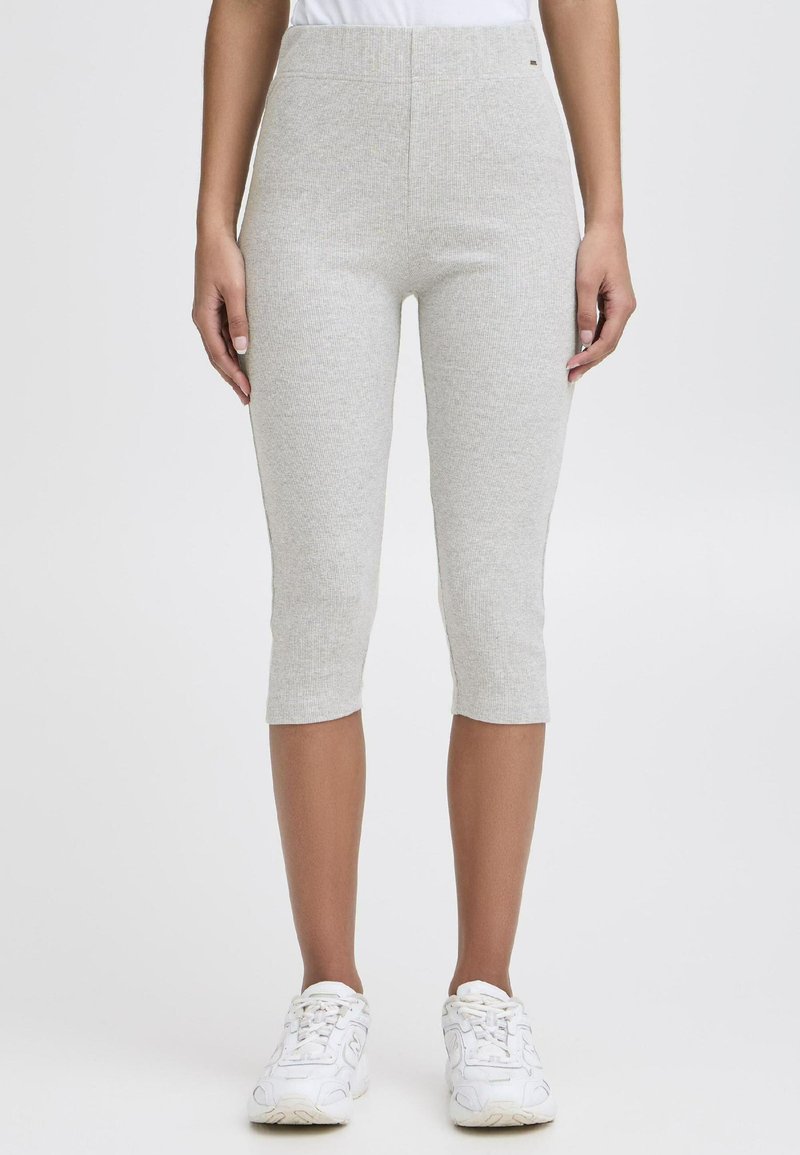 Personne portant des leggings cropped à côtes gris clair et des baskets blanches, se tenant devant un fond blanc uni.