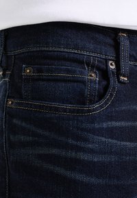 Tmavě modré džínové kalhoty s jemným vyblednutím, s malou zadní kapsou, zlatým šitím a kovovými detaily na knoflíku.