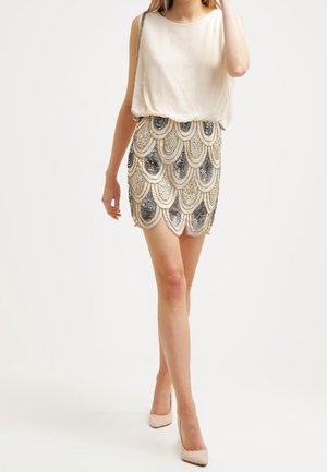 Femme portant un chemisier blanc sans manches et une jupe courte perlée avec des motifs en sequins festonnés, associée à des talons hauts beiges.