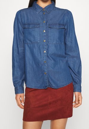 Femme portant une chemise en denim bleue à boutons avec deux poches poitrine et manches longues, associée à une mini-jupe en daim rouge.