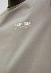 Gaiši pelēka T-krekls, izgatavots no mīksta auduma, ar baltiem drukātiem uzrakstiem "UNIVERSAL" un "Project Core" kontrastējošā dizainā.