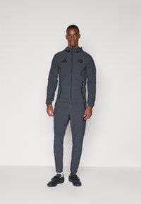 Donkergrijs Adidas trainingspak met ritsvest en slim fit broek. Voorzien van logo op de borst, zakken met ritssluiting en zwarte sportschoenen.