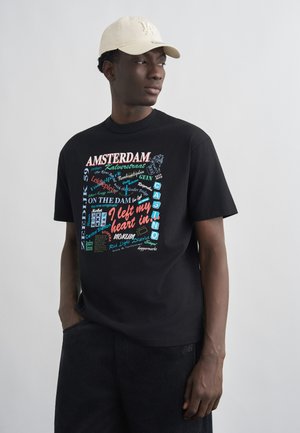 Jaunas vyras dėvi smėlio spalvos kepurę ir juodą marškinėlį su spalvingais Amsterdamo tematikos užrašais ir grafika, stovintis su viena ranka kišenėje.