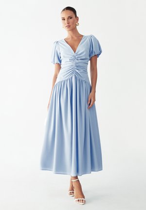 Woman wearing a light blue midi dress with puffed sleeves, ruched bodice, and white ankle-strap heels, standing against a white background.