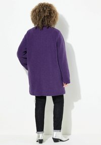 Abrigo largo de terciopelo púrpura, con un ajuste relajado y cuello redondo. Lleva puesto sobre jeans negros y botas de tobillo plateadas con tacón ancho.