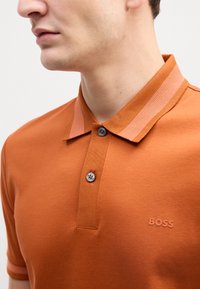 Poloko s krátkym rukávom v spálenej oranžovej bavne, s rebrovaným golierom v svetlejšom odtieni, s trojicou gombíkov a reliéfnym logom.