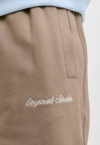 Bež hlace s mehkimi teksturami, ki imajo stranske žepke in vezeno besedilo "Originals Studio" v beli barvi na levi strani.