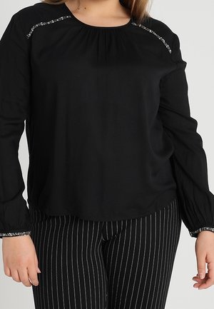 Kobieta ubrana w czarną bluzkę z długim rękawem z cekinowymi detalami na ramionach i mankietach, w zestawieniu z czarnymi spodniami w prążki.