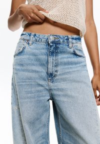 Jasnoniebieskie jeansy z wysokim stanem i zwężaną nogawką, wykonane z denimu. Wśród cech znajdują się zapięcie na guzik, kieszenie na przodzie oraz delikatne przetarcia.