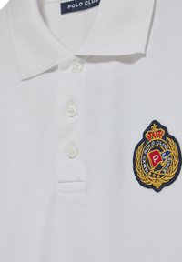 Vit polo shirt gjord av strukturerad tyg med en knäppning med tre knappar och en broderad emblem i blått, rött och guld på vänster bröst.