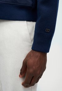 Ναυτικό πλεχτό πουλόβερ με ριγέ μανσέτες, συνδυασμένο με ανοιχτό γκρι παντελόνι. Διαθέτει μια μικρή επιγραφή λογότυπου στο μανίκι, αναδεικνύοντας την υφή του υφάσματος.